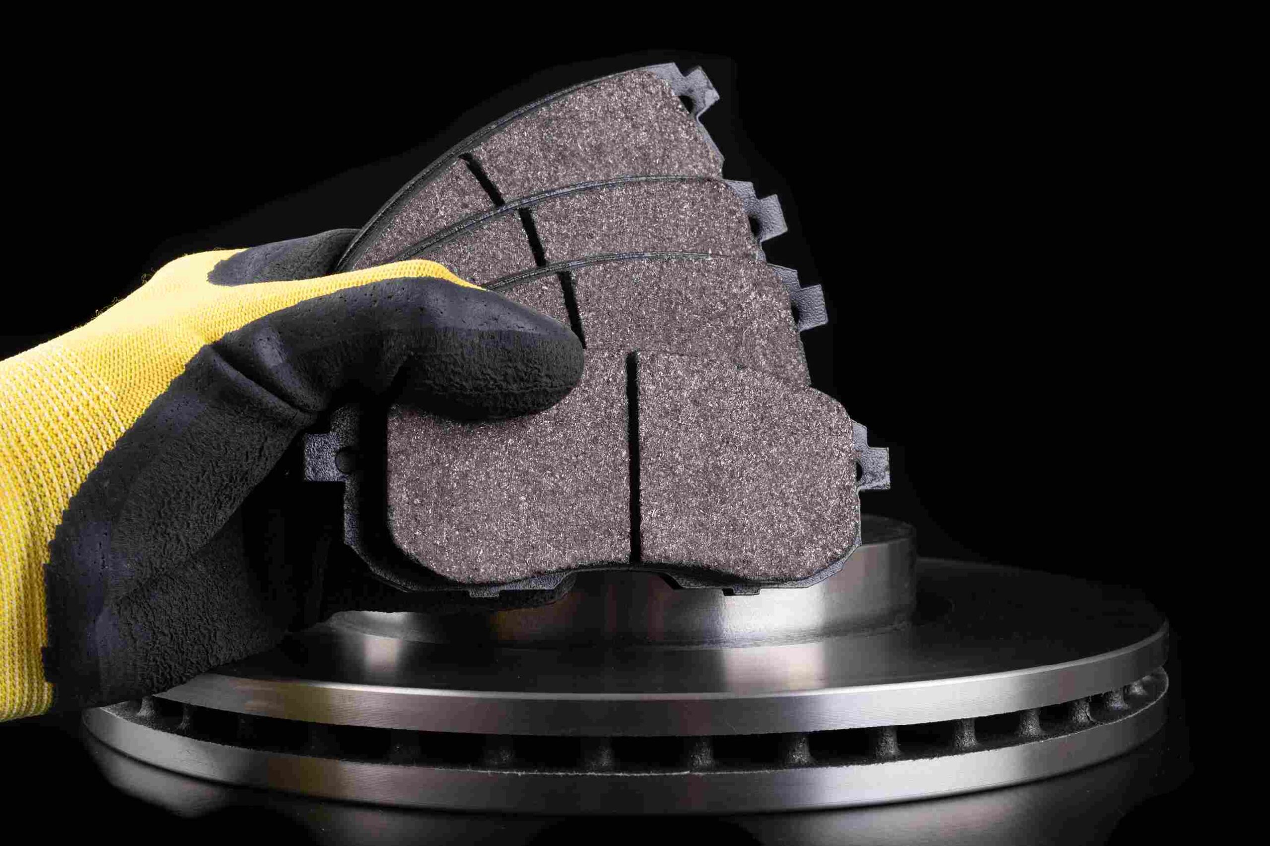 Gloved hand holding brake pads in front of a metal brake rotor on a dark background