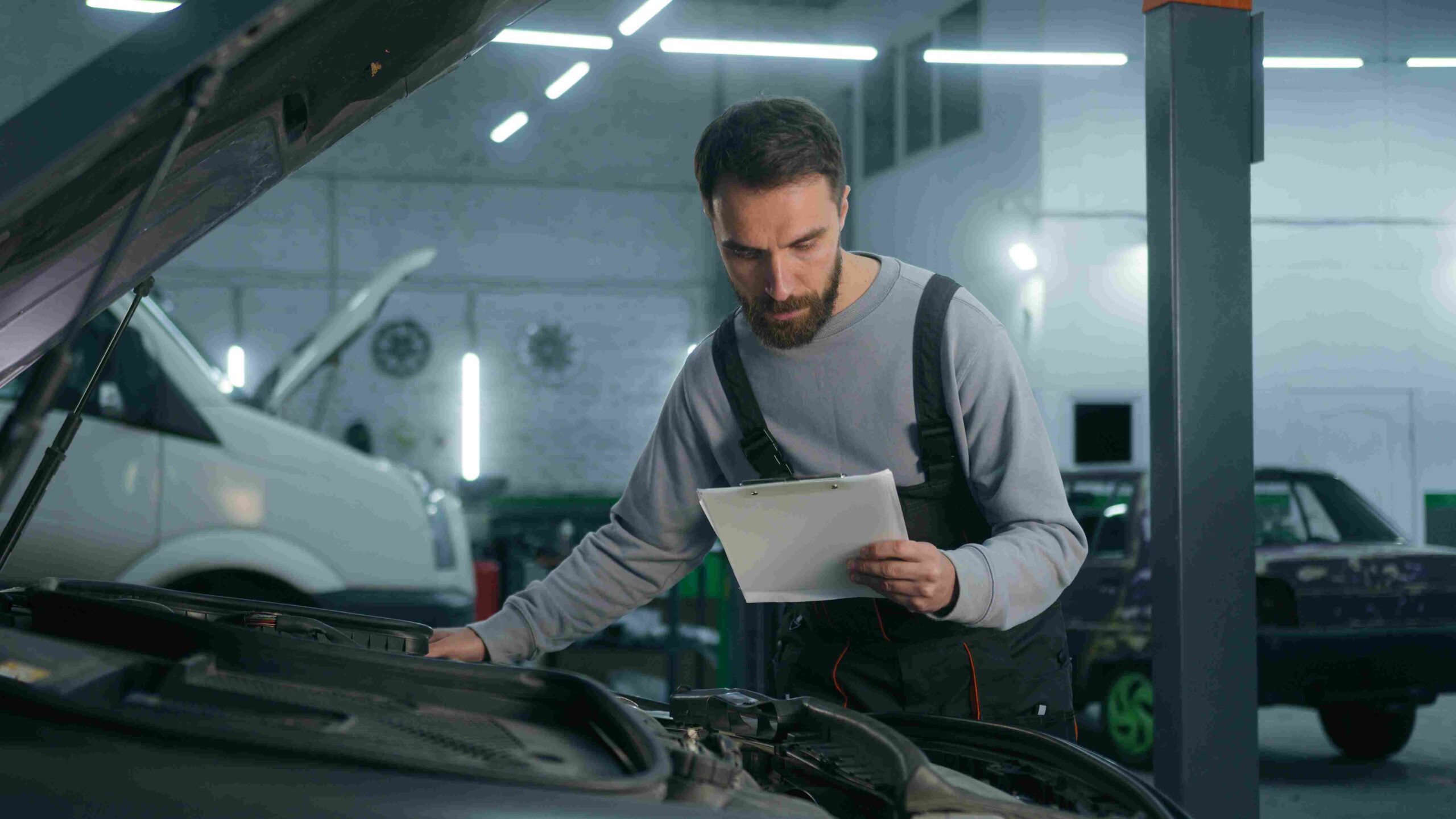 A mechanic checking a car engine while holding a clipboard in a garage