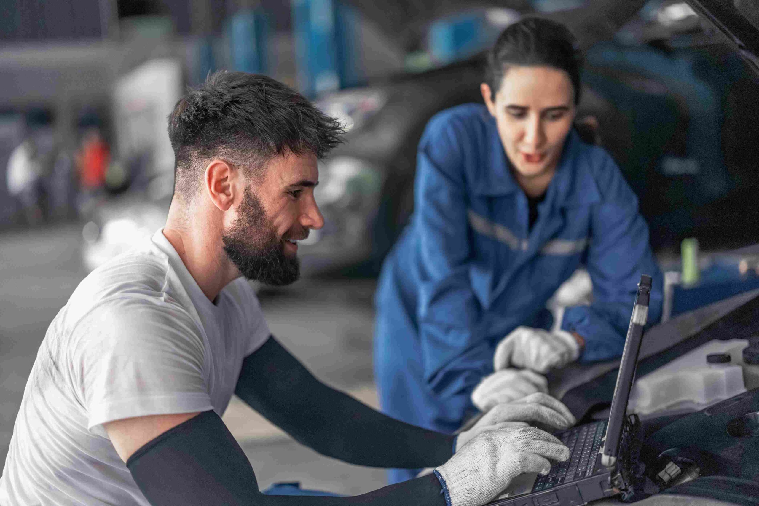 Two mechanics working on a car engine while checking data on a laptop