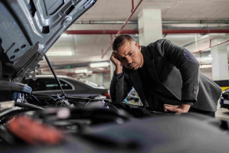 A frustrated man inspecting a car engine under an open hood in a parking garage