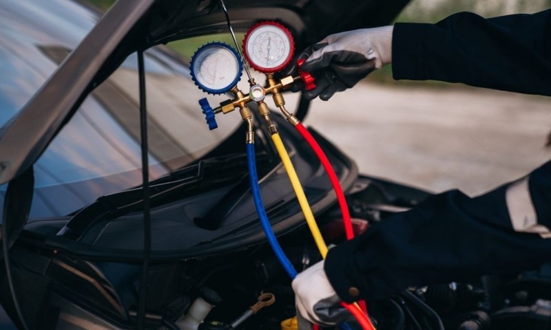 A mechanic in gloves holds a manifold gauge with red and blue hoses, connected to a car's AC system